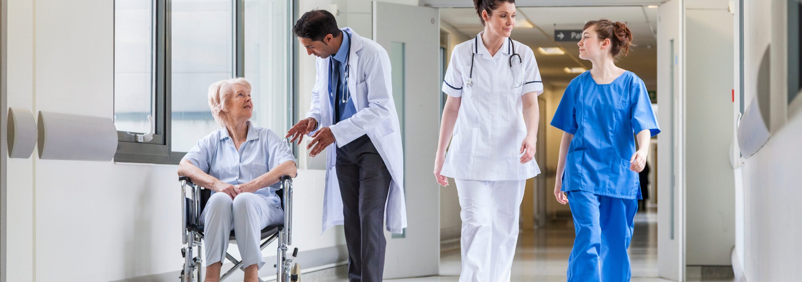 Doctors & nurse in hospital corridor with senior female patient in wheel chair with male Asian doctor